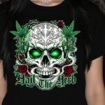 Gothic illustration of a detailed skull with electric green glowing eyes, surrounded by cannabis leaves and red roses on a black background. Gothic font text below reads: Hail The Herb. High-quality PNG for digital printing and merchandise.